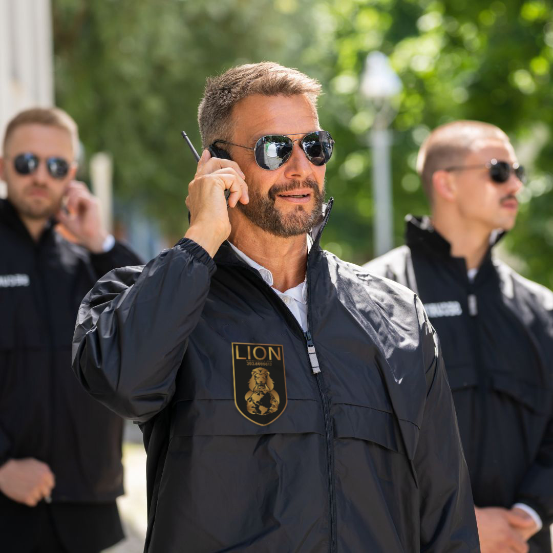 Guardia con occhiali da sole parla alla radio, giacca nera con stemma dorato LION, due colleghi dietro