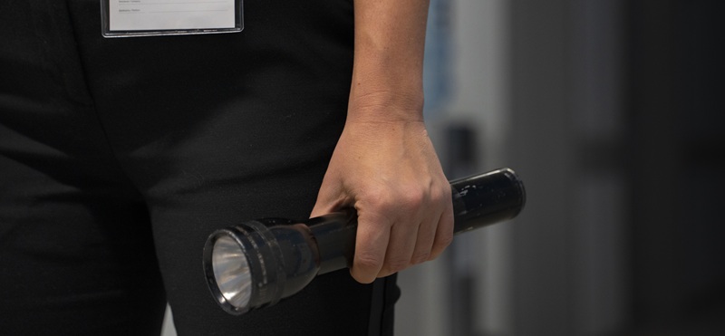 Mano di un addetto in divisa nera con badge che impugna una torcia metallica nera accesa in un corridoio