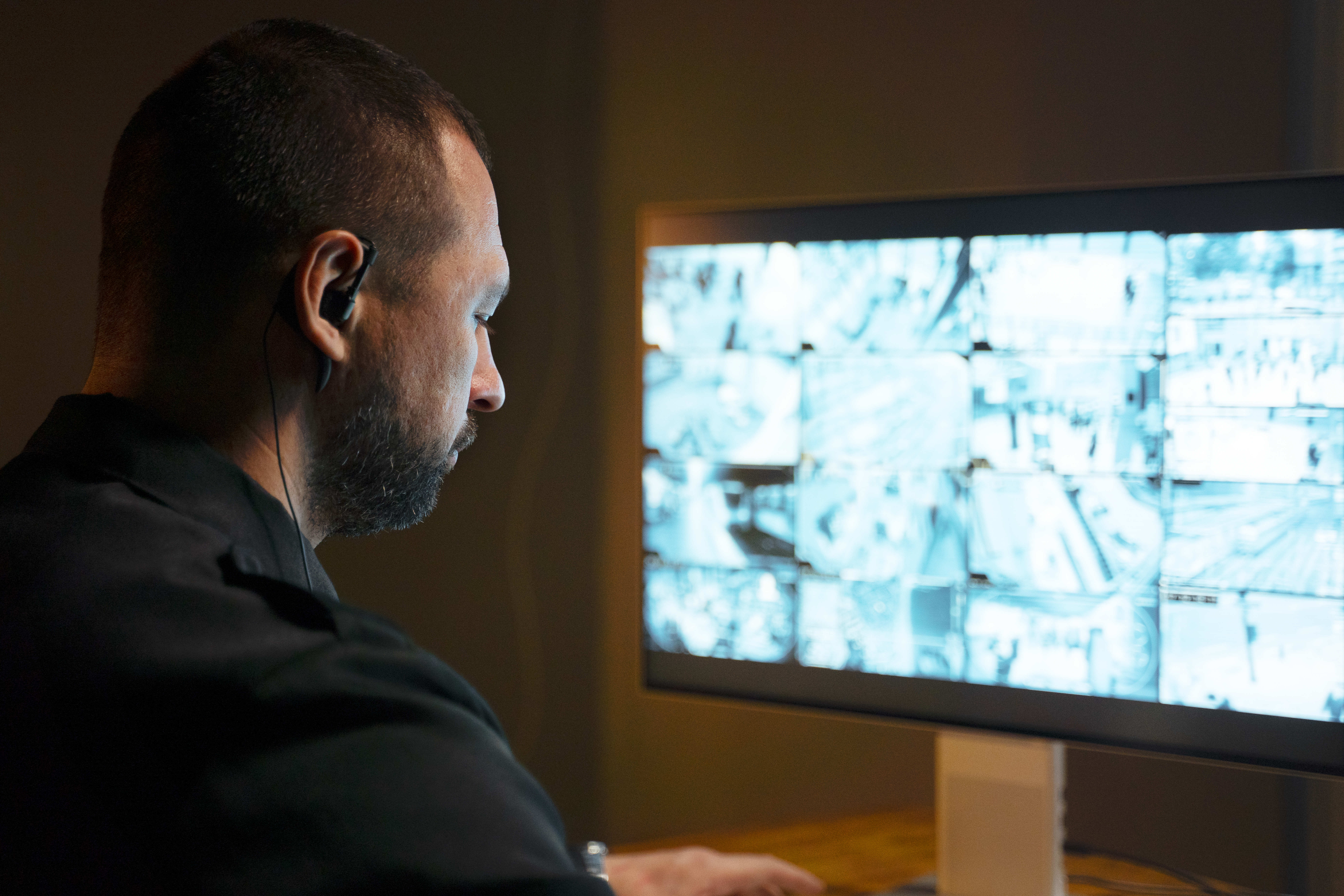 Operatore con auricolare e camicia nera osserva un monitor con griglia di riprese delle telecamere di sorveglianza